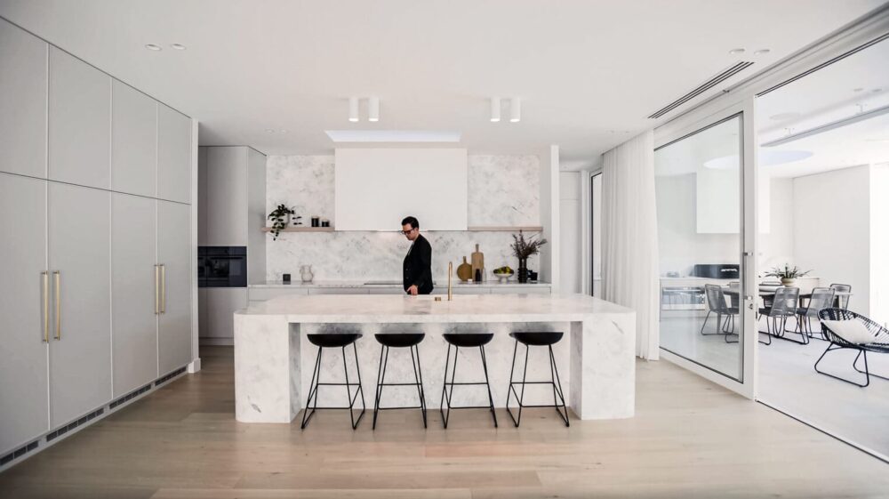 Modern kitchen in a real estate video features marble countertops, sleek grey cabinets, and a man in business attire by the island, showcasing luxury.