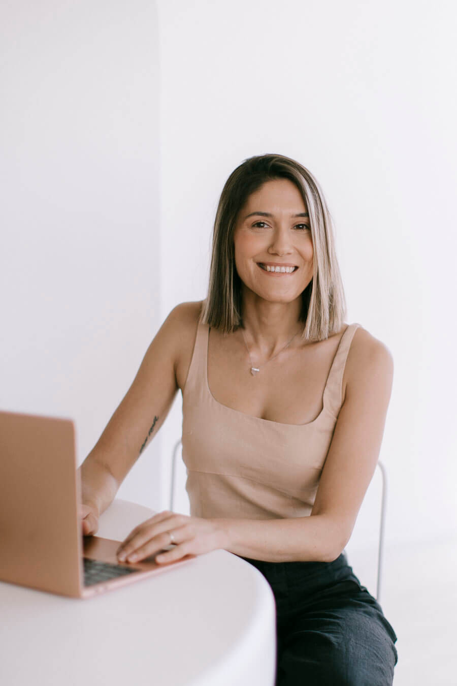 Sera Wilkie, Head of Marketing at DreamCube Productions in Melbourne, sits at a white table with her laptop, smiling warmly at the camera. She projects a professional yet friendly image, dressed in a sleeveless top and black pants, in a bright and minimalistic setting.
