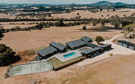 A stunning example of aerial videography showcasing an expansive, modern estate constructed by APC Build, with a series of dark-roofed structures, a neatly kept tennis court, and a pristine swimming pool. The property stands out amidst the broad, open farmlands with scattered trees, capturing the essence of upscale rural living.
