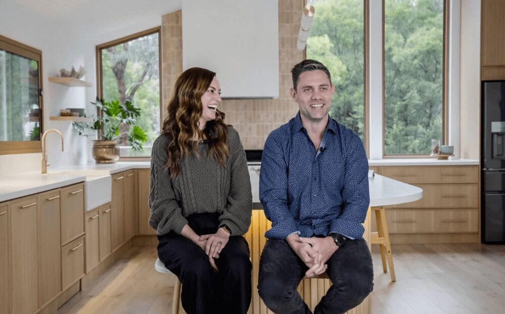 The image captures a cheerful moment from a client testimonial video for Demardi Builders, showcasing a man and a woman seated comfortably in a bright, modern kitchen. The woman, wearing a cozy green sweater and black pants, laughs heartily, turning towards the man who is smartly dressed in a dotted blue shirt and black jeans. He sits with an easy, confident posture, hands clasped and smiling broadly. Behind them, the kitchen exudes warmth with its light wooden cabinets, fresh greenery, and a backdrop of lush trees visible through the large window, illustrating the inviting and harmonious living spaces crafted by Demardi Builders.