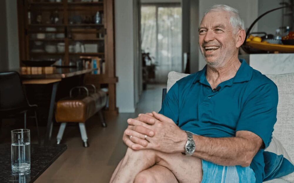The image showcases a joyful senior man from a client testimonial video for Eclipse Tint, comfortably seated and relaxed in a casual setting. He is dressed in a teal polo shirt and light shorts, his legs crossed and hands clasped over his knee. With a silver watch on his wrist, he shares a candid laugh, embodying contentment and ease.