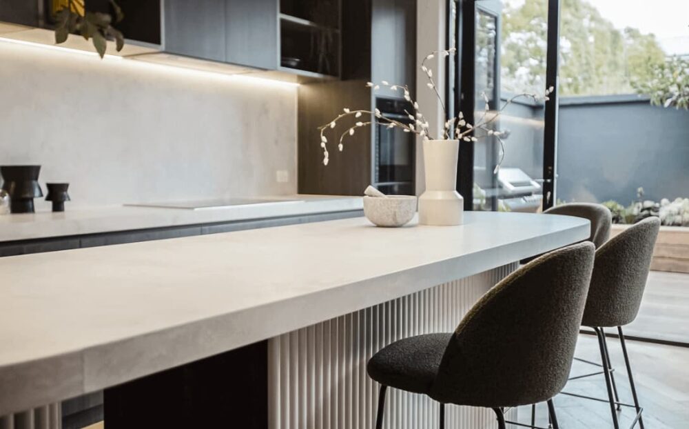 Harrods Build Renovation Video: Modern kitchen with minimalist design, long island, white stone countertop, dark stools.