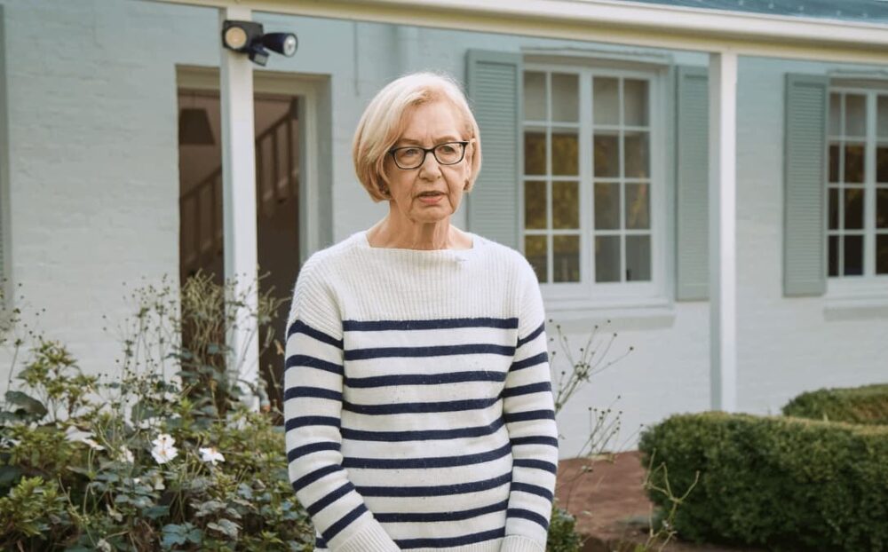 A serene elderly woman stands in her garden, dressed in a casual navy and white striped sweater. She looks directly at the camera with a calm, reflective expression. Behind her is the exterior of her home, freshly painted by 