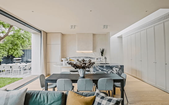 A spacious, light-filled dining area with elegant, modern furniture opens up to a lush outdoor patio, part of a project showcase video by Zeid Projects. The room features a striking marble kitchen island and matching backsplash, harmonizing with the natural wooden flooring and cabinetry.