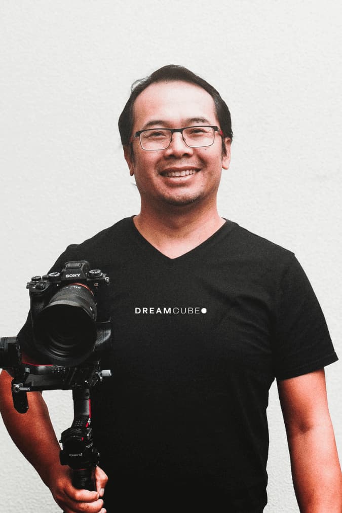 A portrait of Dao Kao, a videographer at DreamCube Productions. He is smiling and wearing glasses, a black T-shirt with the DreamCube logo, and holding a Sony camera mounted on a handheld gimbal stabilizer. The background is a plain, light-colored wall that contrasts with the black of his T-shirt and the camera equipment, highlighting his professional appearance and friendly demeanor.